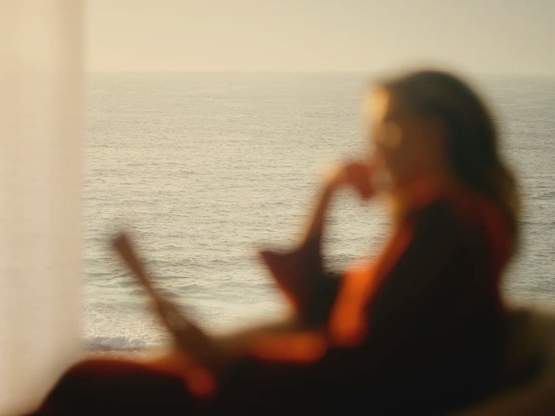 A woman seated by the sea at golden hour, softly out of focus, looking out toward the horizon