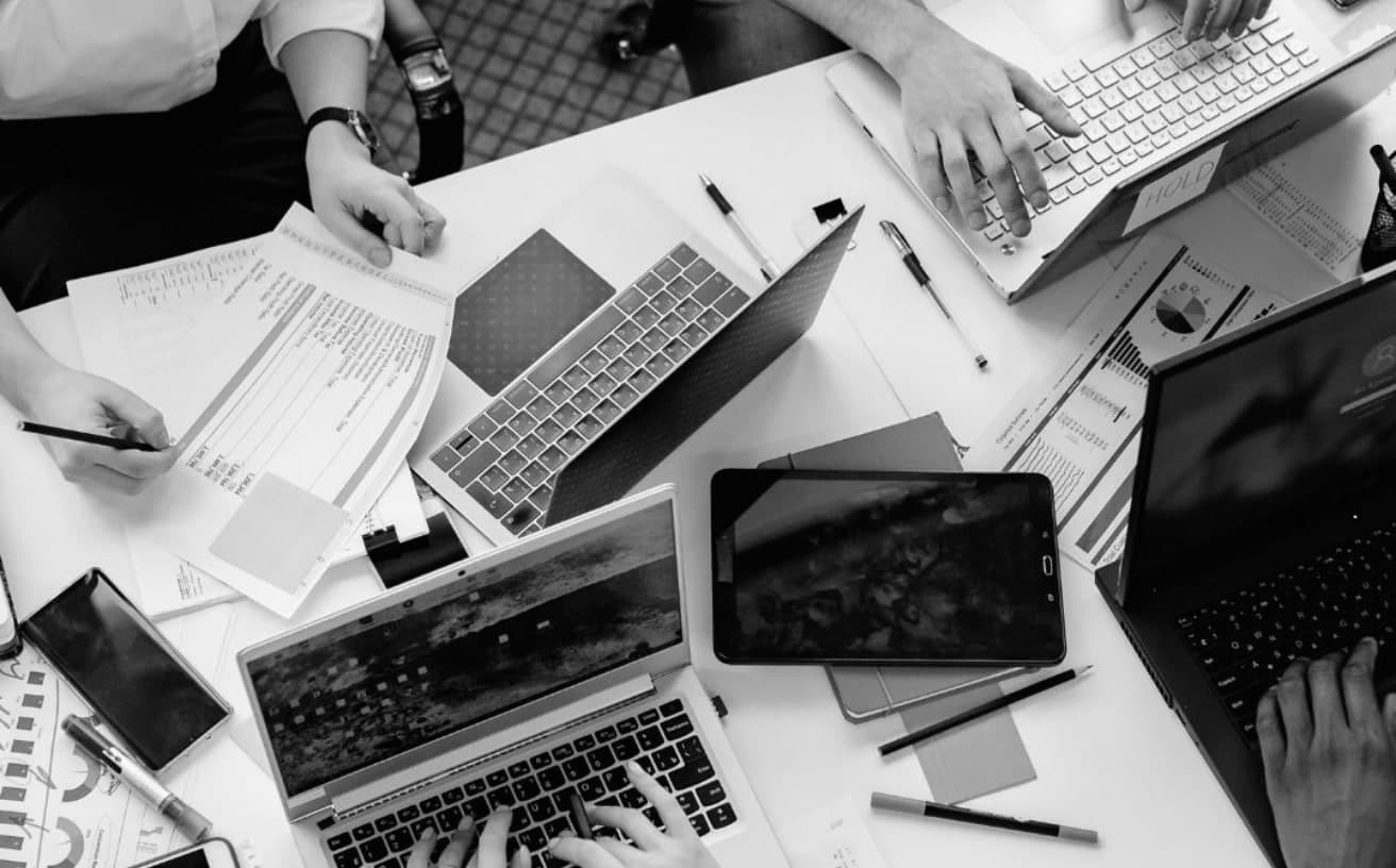 An overhead view of a team working across laptops, a tablet and printed reports covered in charts, on a shared white desk
