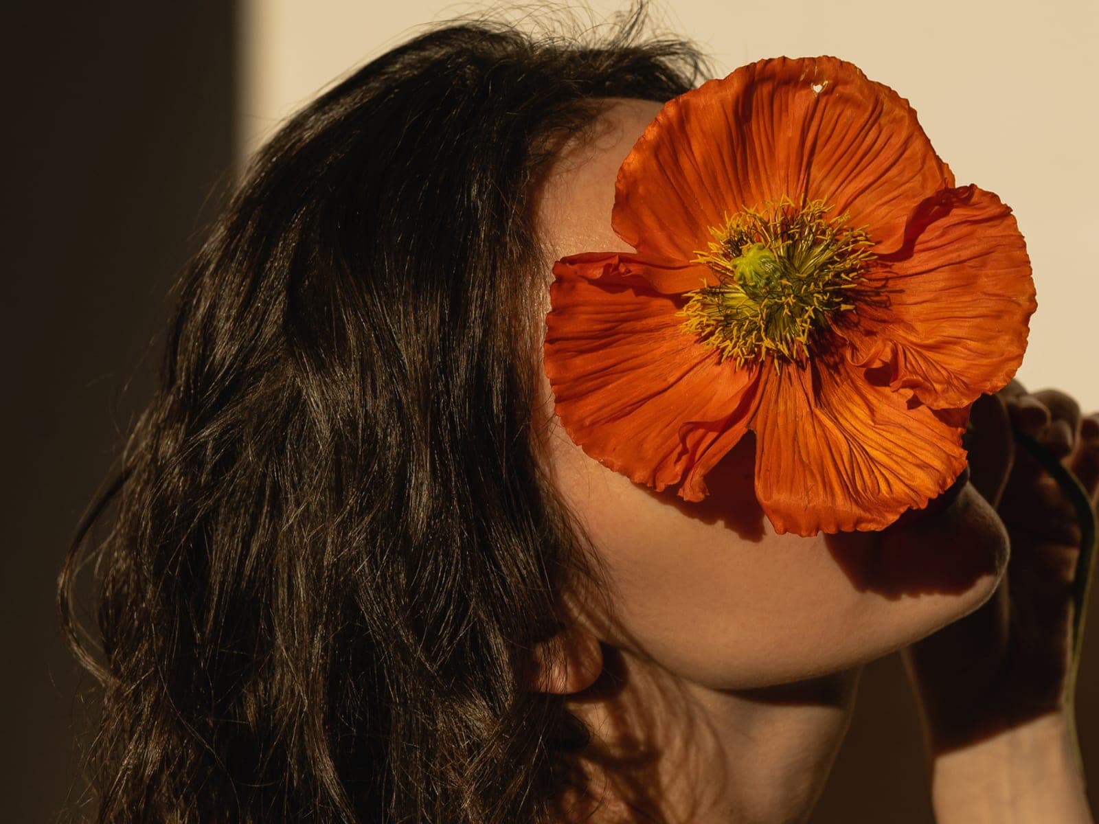 A woman holding a single poppy flower in front of her eyes, lit in warm sunlight