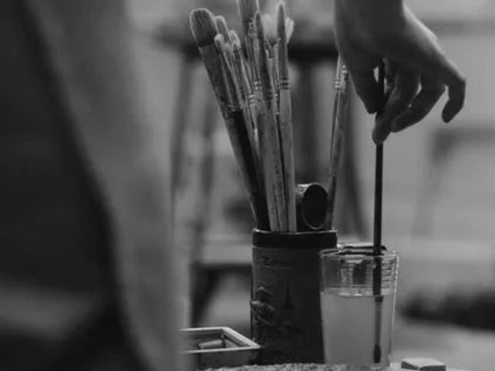 A hand reaching for a paintbrush among a jar of brushes, in black and white