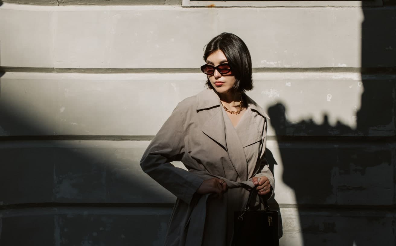 A woman in a beige trench coat and sunglasses standing against a sunlit stone wall, sharp diagonal shadows cast across the scene