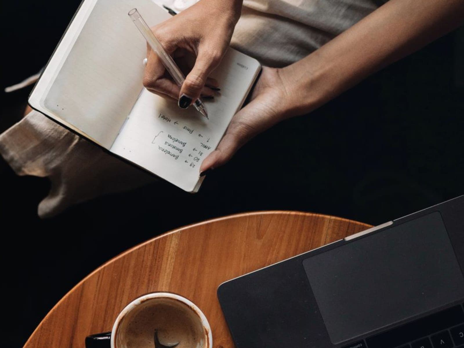 A hand writing a route of destinations in a notebook beside a laptop and coffee