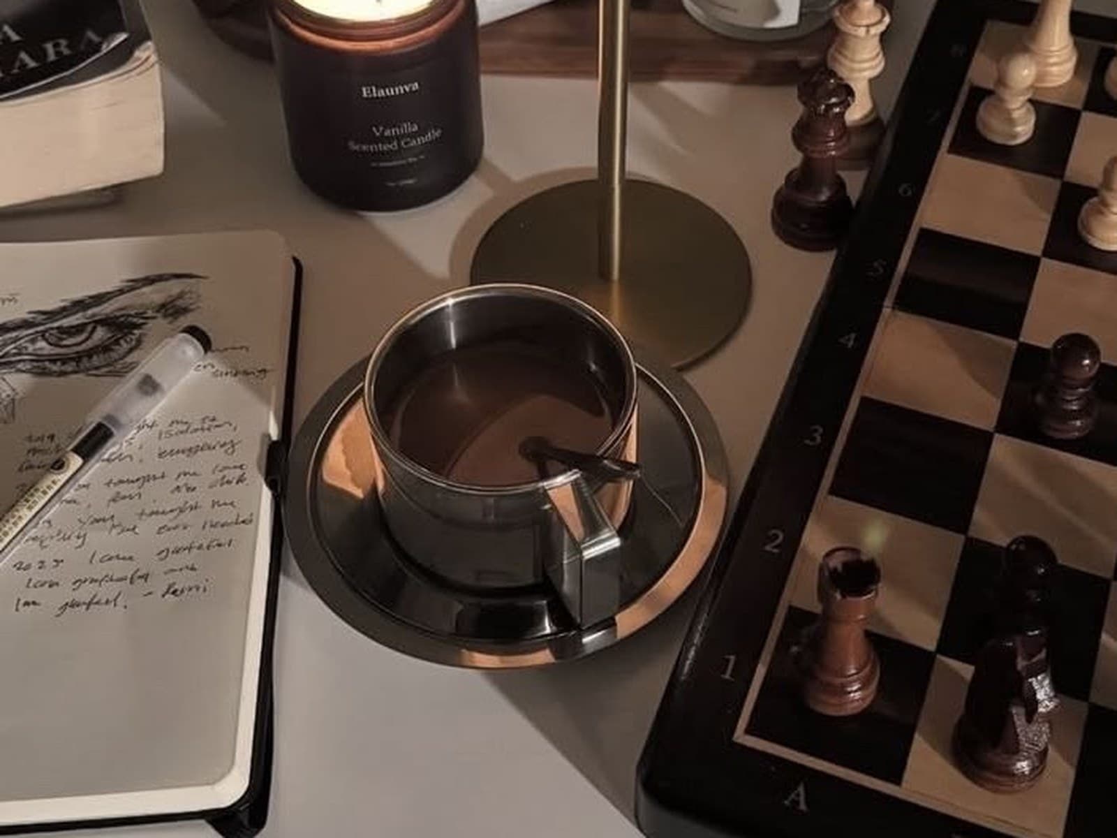 A lit candle, journal and coffee cup beside a chessboard mid-game on a desk