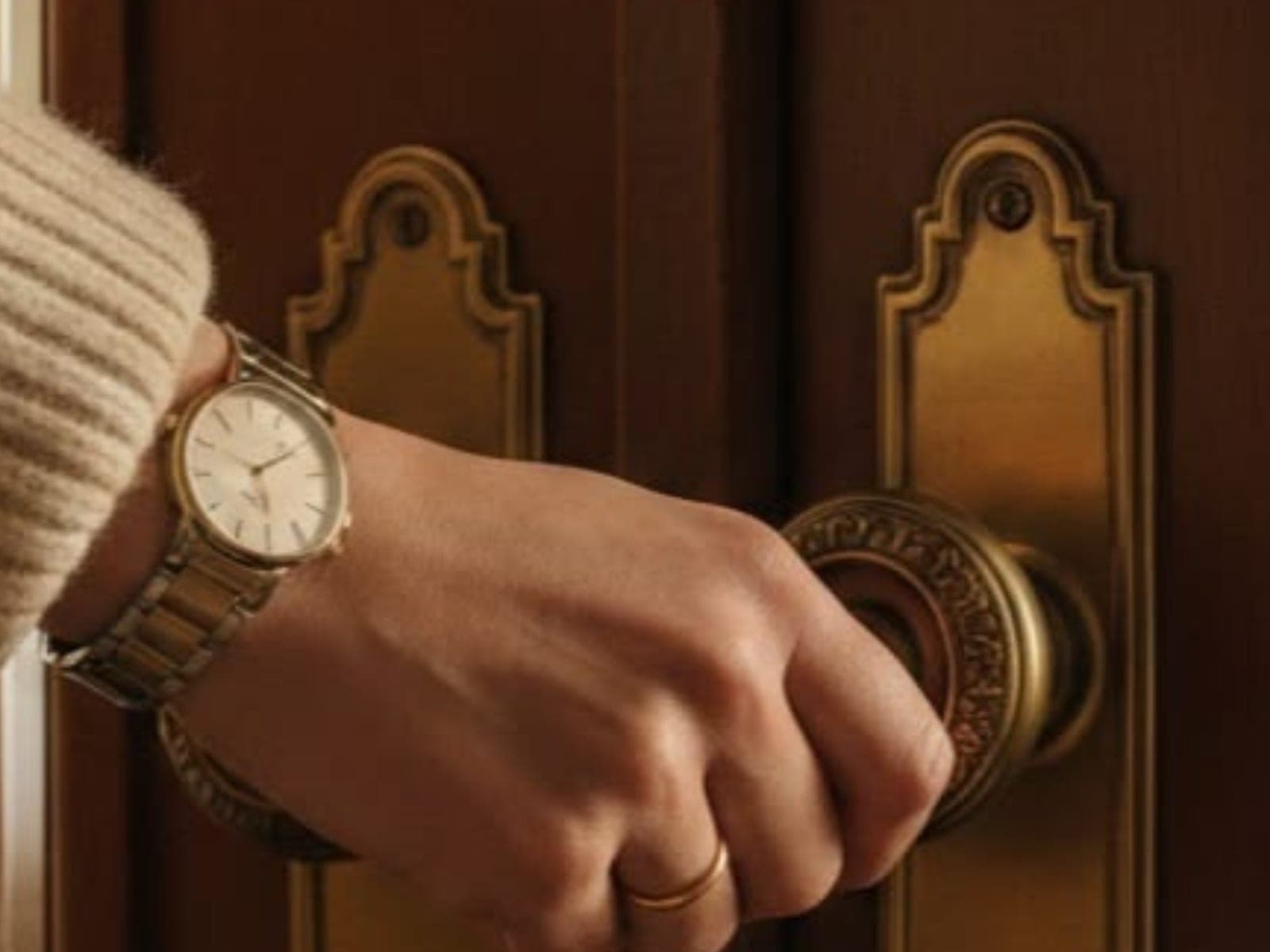 A hand turning an ornate brass door handle, wearing a wristwatch
